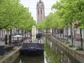 A calm river with boats and green trees on both sides, with a church tower in the background,