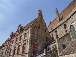 Complex of old brick buildings with many windows and red accents under a blue sky, Bruges,