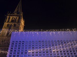 An imposing church tower and a modern building side by side, with festive lighting at night, Ahaus,