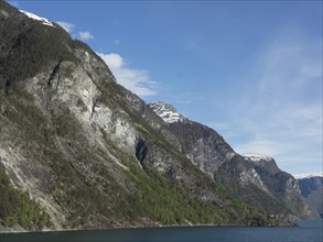 High, snow-covered mountains with green slopes and a lake below under a clear blue sky, Flam,