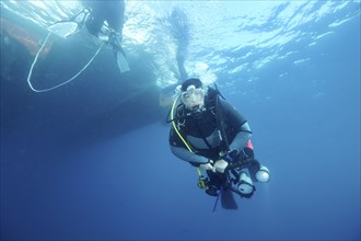 A diver with a camera swims underwater next to a boat on the surface. Dive site Marine reserve Cap