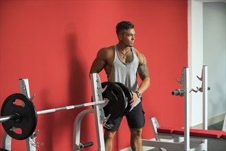 In gym. Tattooed young athlete standing near barbell