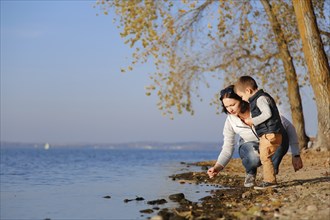 A woman and a child are by a lake on an autumn day, picking something up from the shore while trees