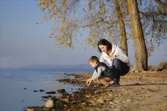 A mother and her child explore the shore of a calm lake during autumn surrounded by trees with