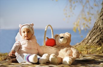 Baby sitting on a blanket outdoors with a teddy bear and basket containing a pumpkin on a sunny