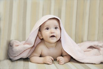 Baby under a pink towel looking up while lying on a striped beige surface, Belarus.Minsk
