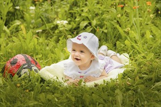 A smiling baby laying on the grass with a white hat, surrounded by flowers and greenery in the