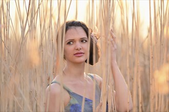 A young woman with brown hair stands thoughtfully amidst reeds with soft sunlight, Bavaria