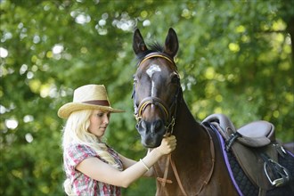 Woman (cowgirl) in a straw hat and plaid shirt stands beside a polish arabian horse on a field,