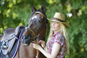 Woman (cowgirl) in a straw hat and plaid shirt stands beside a polish arabian horse on a field,