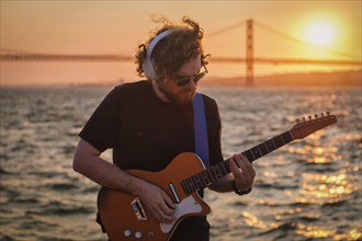 Hipster street musician in black playing electric guitar in street outdoors on sunset