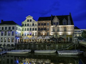 Riverbank at night with illuminated buildings and restaurants. Several boats are moored on the