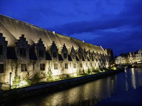 Long historic buildings on the riverbank illuminated at night under a deep blue sky, Ghent,