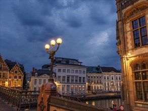 Historic buildings along a river at dusk, illuminated lanterns on the bridge, Ghent, Belgium,
