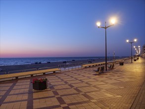 An empty promenade at dusk, illuminated by lanterns, the sea in the background, a peaceful and