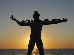 A statue with birds on its shoulders stands in the sunset, with the sea in the background, showing