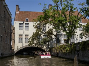 Boat sailing through a narrow canal, past old buildings and trees under a sunny sky, Bruges,