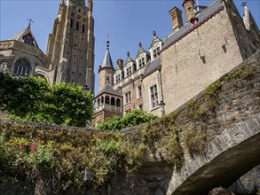 Historic buildings and church tower, surrounded by plants, near a bridge under blue sky, Bruges,