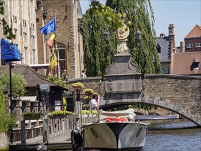A boat under a bridge near historic buildings, decorated with flags and flowers, Bruges, Belgium,