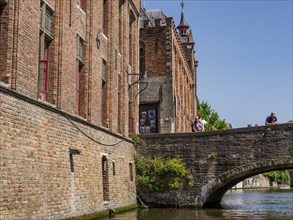 Tourists on an old stone bridge next to a brick building over a canal, Bruges, Belgium, Europe