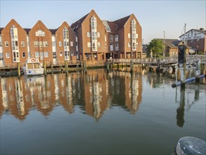 Brick buildings and jetties are reflected in the calm waters of the canal, a few boats can be seen,