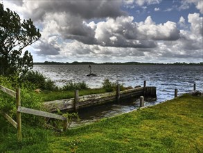 Lake view with jetty and lush shore grass under a dramatic sky with dense clouds, Maasholm,