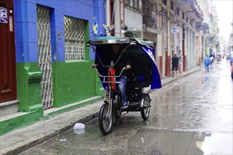 Havana, Cuba, Central America, man riding a cycle rickshaw on a wet road in an urban environment,
