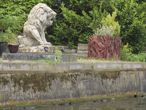 Stone statue of a lion on an overgrown and watered stone bank surrounded by nature, Leiden,