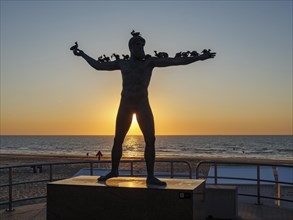 Sculpture on the beach at sunset with bird figures on the arms, silhouette against the light, sea