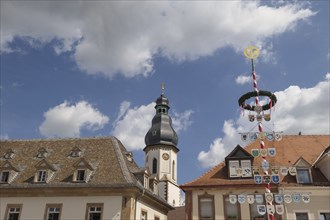 Historic buildings and a church tower decorated with coats of arms and a maypole under a cloudy
