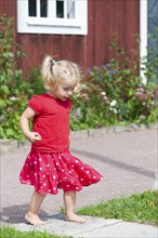 Little blond girl with pigtails in a red summer dress in the garden in front of a typical red