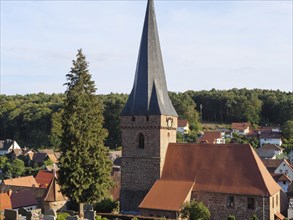 Pointed church tower with red roof and surrounding village houses, surrounded by trees under a blue