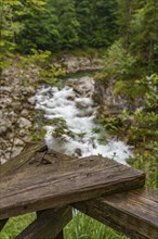 A wooden railing in the foreground with a view of a flowing mountain stream and rocks, surrounded