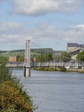 Close-up of a bridge crossing a river with houses and green hills in the background under a cloudy