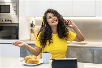 Latin happy young woman dancing while listening to music in the morning in the kitchen at home