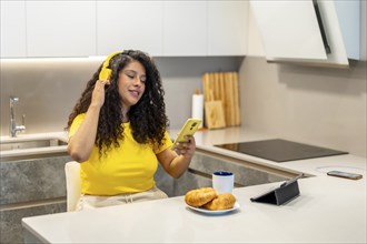 Latin young woman listening to music while eating breakfast in the kitchen at home