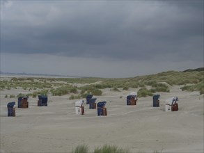 Scattered beach chairs on a sandy beach between grass and dunes under threatening clouds, Juist,