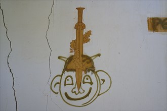 Graffiti of a face with a sword can be seen on a cracked white wall of an abandoned building, Lost