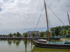 A sailing boat is moored in a quiet harbour, surrounded by buildings and trees under a cloudy sky,