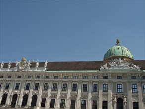 White baroque building with green domed roof and statues on the long façade under a blue sky,