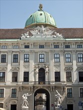 Historic baroque building with green domed roof, archway and statues under a clear sky, Vienna,
