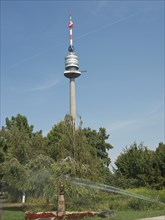 A tall television tower in a park with trees and a running irrigation system, Vienna, Austria,