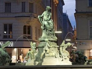 Illuminated fountain with a statue and several figures in an evening city environment, Vienna,