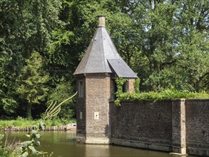 Brick round tower with grey pointed roof on a moat, surrounded by green summer trees and historic