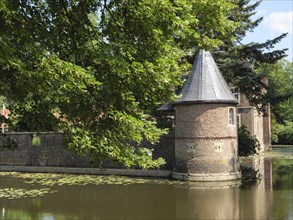 Brick round tower with grey roof, surrounded by a moat and summer greenery, view of historic