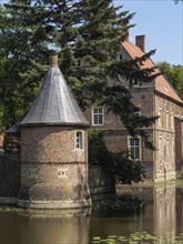 Brick round tower with pointed roof next to a moat, surrounded by summer trees and historic