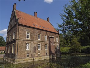 Old brick house with red roof on the edge of a pond, surrounded by green trees in summer, ochtrup,