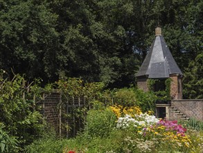 Lush garden with an old brick tower and blooming flower beds surrounded by tall trees, ochtrup,