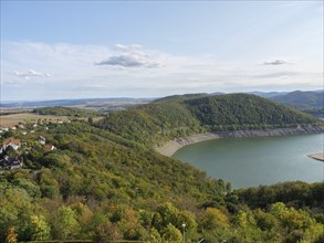 A tranquil lake is nestled in a landscape surrounded by green hills under a blue sky, Waldeck,