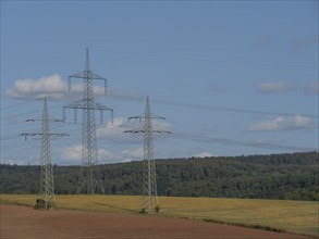 Electricity pylons over wide agricultural fields with a forest in the background, Waldeck, Hesse,